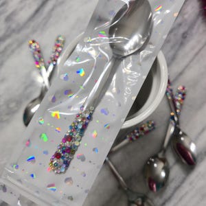 May include: A stainless steel spoon with a handle embellished with multicolored rhinestones, encased in a clear plastic sleeve featuring a holographic heart pattern. Additional similar spoons with rhinestone-decorated handles are visible nearby.