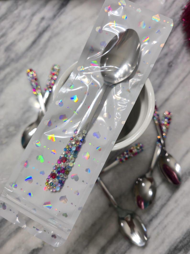 May include: A stainless steel spoon with a handle embellished with multicolored rhinestones, encased in a clear plastic sleeve featuring a holographic heart pattern. Additional similar spoons with rhinestone-decorated handles are visible nearby.