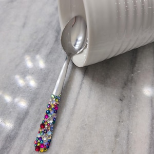 May include: A silver-toned metal spoon with a handle embellished with a dense array of multicolored, faceted rhinestones in shades of red, blue, yellow, and clear. The spoon is positioned partially inside a white, ribbed ceramic mug.
