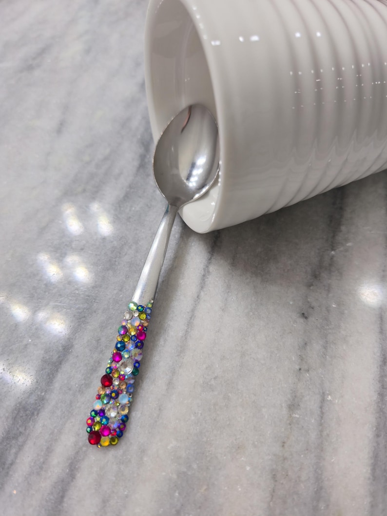 May include: A silver-toned metal spoon with a handle embellished with a dense array of multicolored, faceted rhinestones in shades of red, blue, yellow, and clear. The spoon is positioned partially inside a white, ribbed ceramic mug.