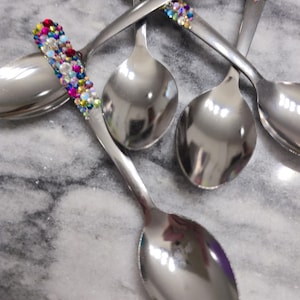 May include: A set of four silver-toned metal spoons with handles embellished with a dense arrangement of small, multi-colored rhinestones in shades of blue, pink, yellow, and clear, creating a vibrant, reflective, and textured decorative finish.