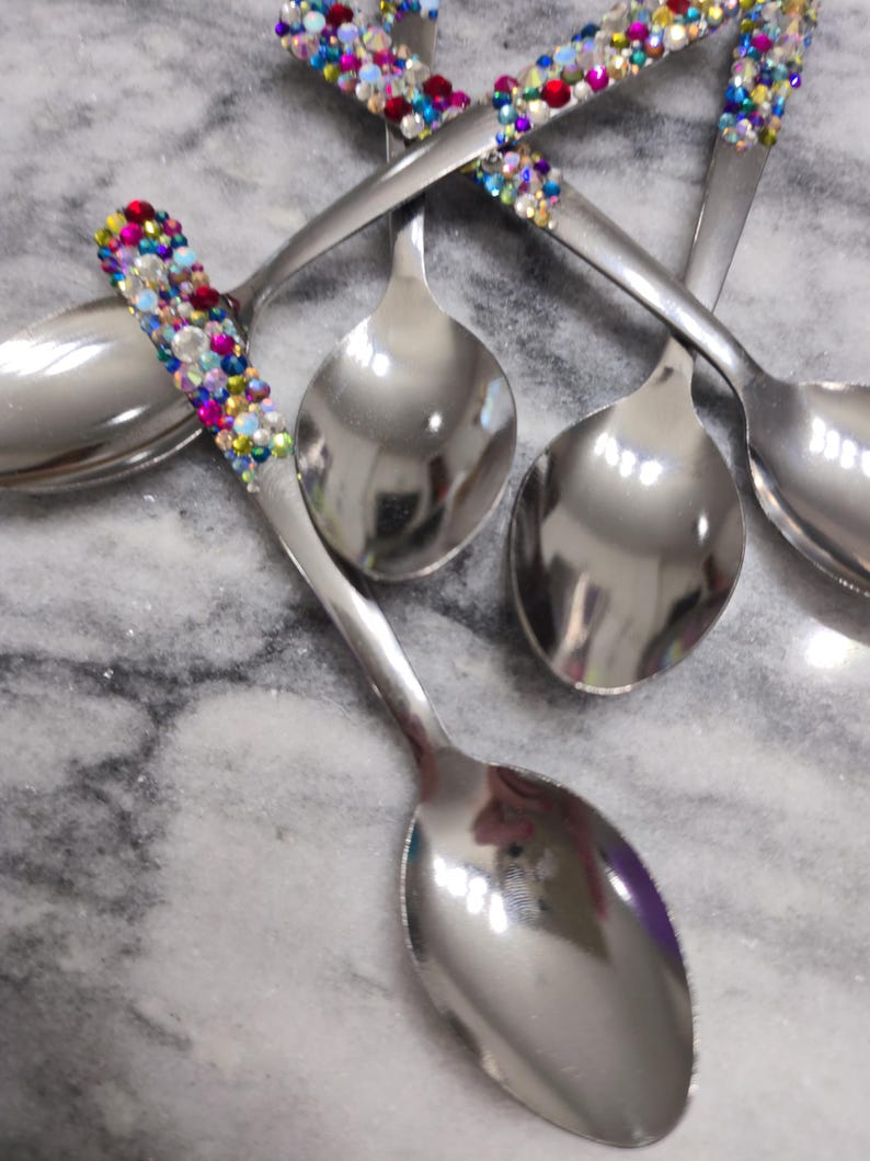 May include: A set of four silver-toned metal spoons with handles embellished with a dense arrangement of small, multi-colored rhinestones in shades of blue, pink, yellow, and clear, creating a vibrant, reflective, and textured decorative finish.