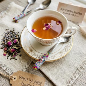May include: A white porcelain teacup and saucer with gold trim containing tea with pink rose petals. Three silver spoons feature handles decorated with multicolored rainbow gems. A brown tag reads SPARKLE TEA SPOON - RAINBOW GEM.