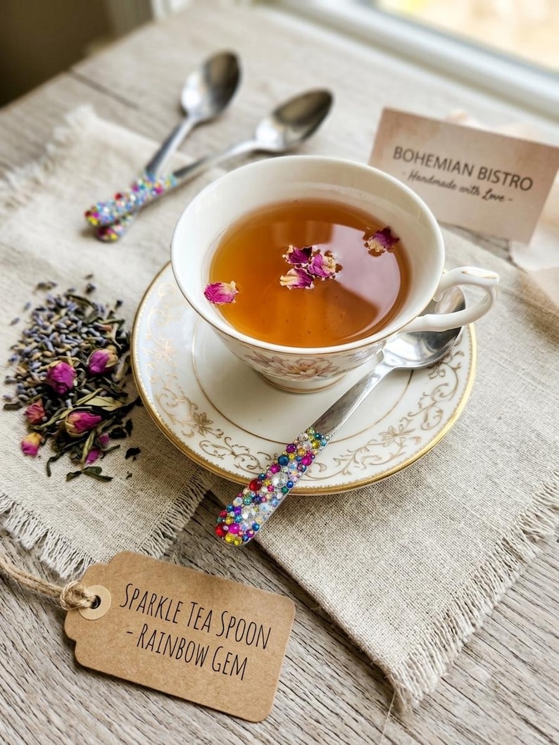 May include: A white porcelain teacup and saucer with gold trim containing tea with pink rose petals. Three silver spoons feature handles decorated with multicolored rainbow gems. A brown tag reads SPARKLE TEA SPOON - RAINBOW GEM.