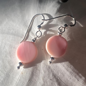 May include: A pair of dangle earrings featuring pale pink, circular beads. The earrings have silver-colored hooks and small, round accents. The beads have a subtle, layered texture. The earrings are displayed on a white, textured surface.