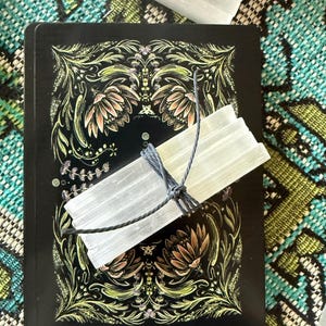 May include: A black tarot card with floral and leaf designs, and a bundle of selenite crystals tied with gray string. The crystals are translucent and rectangular. The background is a patterned teal and green fabric.