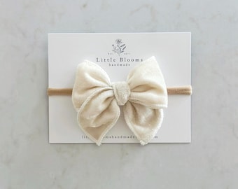 May include: A cream-colored velvet bow headband with a nude elastic band. The headband is on a white card with the text "Little Blooms handmade".