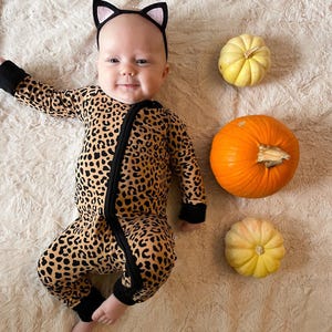 Op de afbeelding: Een baby die een luipaardprint onesie en een zwart kattenoren-hoofdband draagt, liggend op een beige pluche oppervlak met drie kleine pompoenen ernaast.