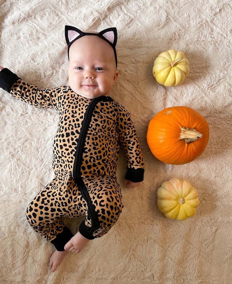 Op de afbeelding: Een baby die een luipaardprint onesie en een zwart kattenoren-hoofdband draagt, liggend op een beige pluche oppervlak met drie kleine pompoenen ernaast.