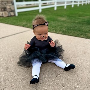 Op de afbeelding: Een baby verkleed als een zwarte kat voor Halloween. De baby draagt een zwarte tutu, een zwart shirt met lange mouwen en een zwart kattenhaarband met oren. De baby heeft zwarte katten snorharen op zijn gezicht geschilderd.