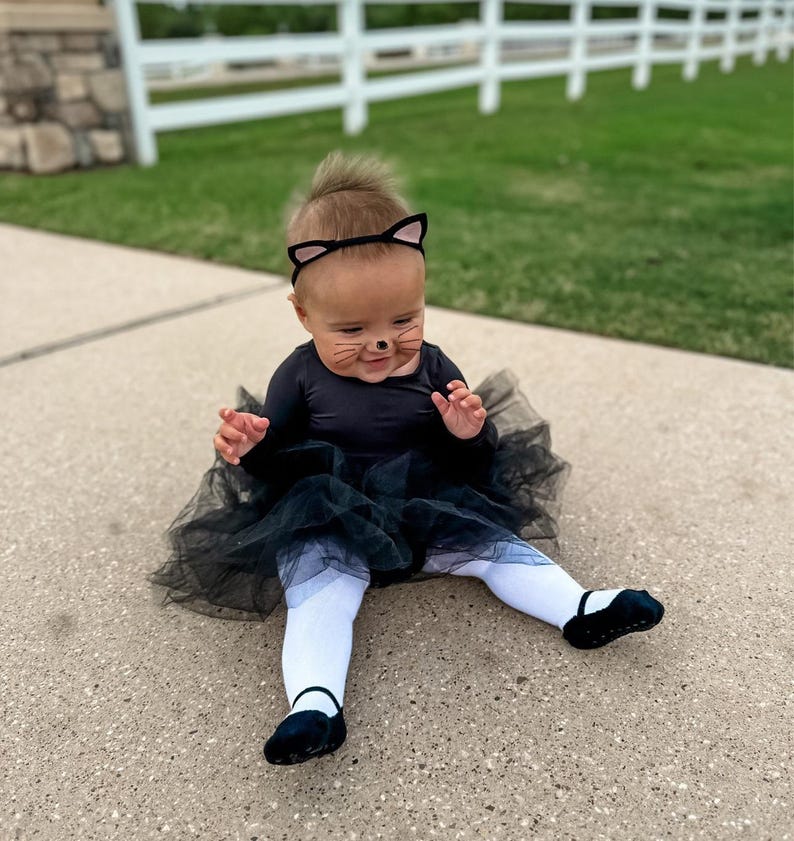 Op de afbeelding: Een baby verkleed als een zwarte kat voor Halloween. De baby draagt een zwarte tutu, een zwart shirt met lange mouwen en een zwart kattenhaarband met oren. De baby heeft zwarte katten snorharen op zijn gezicht geschilderd.