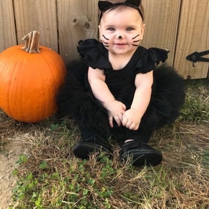 Op de afbeelding: Een baby gekleed in een zwarte tule tutu en een zwart shirt met zwarte kattenoren en gezichtsverf. De baby zit op het gras naast een grote oranje pompoen.