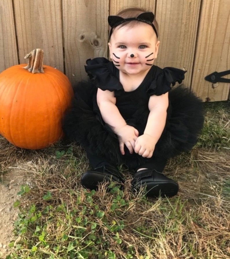 Op de afbeelding: Een baby gekleed in een zwarte tule tutu en een zwart shirt met zwarte kattenoren en gezichtsverf. De baby zit op het gras naast een grote oranje pompoen.