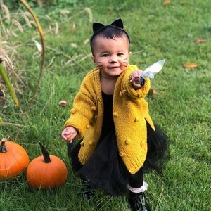 Op de afbeelding: Een jong kind verkleed als kat voor Halloween. Het draagt een gele gebreide cardigan met pompoenen, een zwarte tule rok en zwarte kattenoren. Het kind houdt een witte snoepstok.