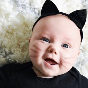 Op de afbeelding: Een baby draagt een zwart kattenhoofdband met zwarte oren en een zwart shirt. De baby heeft bruine katten snorharen op zijn wangen getekend en lacht.