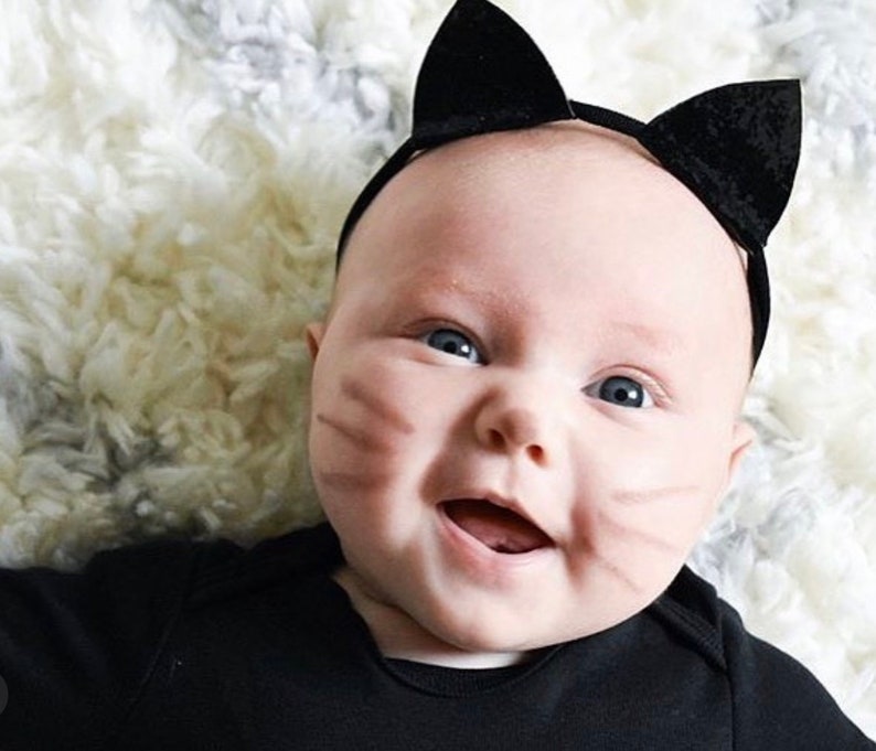 Op de afbeelding: Een baby draagt een zwart kattenhoofdband met zwarte oren en een zwart shirt. De baby heeft bruine katten snorharen op zijn wangen getekend en lacht.