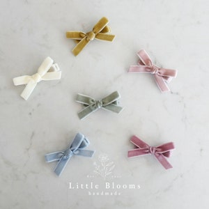 Puede incluir: Seis lazos para el pelo de terciopelo en varios colores, incluyendo azul claro, rosa polvoriento, verde salvia, marfil, amarillo mostaza y rosa. Los lazos están dispuestos sobre una superficie de mármol blanco. El texto "Little Blooms handmade" es visible en la parte inferior de la imagen.