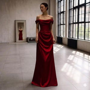 May include: A floor-length, off-the-shoulder, red satin gown. The dress features a fitted bodice and a flowing skirt. The model is standing in a well-lit room with large windows and a mirror reflecting the dress.