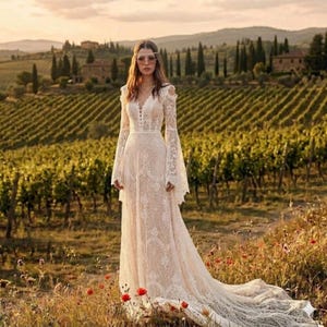 Puede incluir: Vestido de novia de encaje marfil con mangas largas acampanadas y cola. El vestido tiene un escote en V y detalles de botones. El fondo muestra un viñedo y colinas.