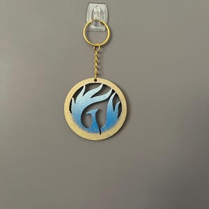 Op de afbeelding: Een ronde sleutelhanger met een goudkleurige ring en ketting. De hanger heeft een blauw en goud phoenix ontwerp in een cirkelvormig frame. De sleutelhanger hangt tegen een grijze muur.