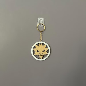 Op de afbeelding: Een goudkleurige sleutelhanger met een cirkelvormige hanger met een leeuwenkopontwerp. De leeuwenkop is goudkleurig, geplaatst in een witte cirkel. De sleutelhanger hangt tegen een grijze muur.