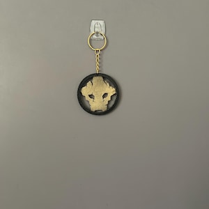 Op de afbeelding: Een goudkleurige, gestileerde dierenhoofd sleutelhanger met een zwarte cirkelvormige rand. De sleutelhanger hangt aan een goudkleurige ring en ketting. De achtergrond is een effen grijze muur.