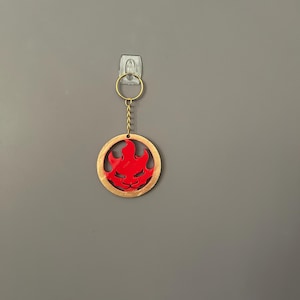 Op de afbeelding: Een rode sleutelhanger in de vorm van een vlam met een goudkleurige rand en een gouden ketting. De sleutelhanger hangt tegen een grijze muur. Het vlamontwerp is een gestileerde weergave.
