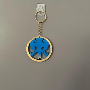 Op de afbeelding: Een blauw en gouden sleutelhanger met een cirkelvormig ontwerp. Het blauwe ontwerp bevindt zich in het midden van de gouden cirkel. De sleutelhanger hangt aan een gouden ring en ketting.