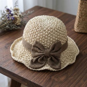 Puede incluir: Un sombrero de paja trenzada beige con una copa redondeada y un ala ondulada. Una banda de tela marrón oscuro rodea la base de la copa, adornada con un lazo prominente y centrado hecho del mismo material marrón oscuro en la parte frontal.