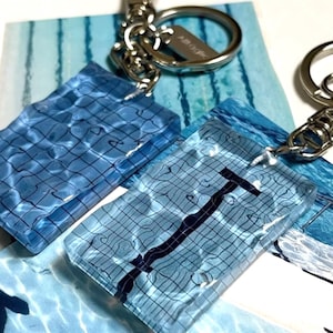Può includere: Portachiavi con ciondoli rettangolari in resina blu. I ciondoli hanno una consistenza simile all'acqua con linee a griglia nere e una linea di corsia di nuoto nera. Sono attaccati portachiavi color argento. Lo sfondo presenta carta in stile acquerello blu e bianco.
