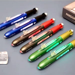 Puede incluir: Un conjunto de bolígrafos con forma de submarino en varios colores, como azul, rojo y verde. Cada bolígrafo tiene un modelo de submarino en miniatura y el número "24" impreso. Los bolígrafos están colocados sobre una superficie gris. Cada bolígrafo mide unos 15 cm.