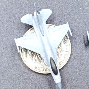 Peut inclure: Un petit avion de chasse miniature en plastique gris clair avec une verrière de cockpit sombre, posé directement sur une pièce de monnaie circulaire. L'appareil présente des ailes en flèche, un fuselage central et un nez pointu moulé.