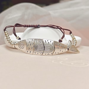 Könnte beinhalten: Ein Armband mit einem zentralen, silberfarbenen Metallanhänger in Fischform, der aus drei beweglichen Segmenten mit eingravierten Details besteht. Der Anhänger ist an einer dunkelbraunen Kordel mit verstellbaren Knoten und silberfarbenen Perlen befestigt.