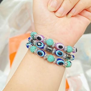 Könnte beinhalten: Ein Set aus drei elastischen Armbändern mit türkisfarbenen Steinperlen, dunkelblauen und violetten Glasperlen mit Nazar-Motiv sowie silberfarbenen Metallzwischenelementen. Die Armbänder sind gestapelt und wechseln sich in den Perlenarten ab.