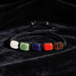 Könnte beinhalten: Ein schwarzes Kordelarmband mit fünf rechteckigen, polierten Steinen in Weiß, Grün, Dunkelblau, Rot und braunem Tigerauge. Das Armband hat ein geflochtenes Makramee-Band mit verstellbaren Schiebeknoten und zwei kleinen schwarzen Perlen an den Enden.