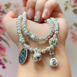Könnte beinhalten: Ein zweireihiges silberfarbenes Perlenarmband mit Metallanhängern. Die Anhänger bestehen aus einem runden gravierten Schild, einer kleinen Glocke und einem Geldsack mit Dollarzeichen. Die Perlen sind gleichmäßig und reflektierend.