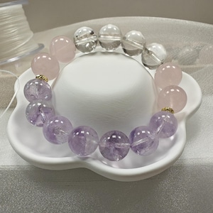 Könnte beinhalten: Ein Armband aus runden, polierten Edelsteinen in den Farben Hellrosa, Transparent und Hellviolett. Die Perlen sind mit kleinen goldfarbenen Metallzwischenstücken zwischen den verschiedenen farbigen Steinabschnitten aufgereiht.