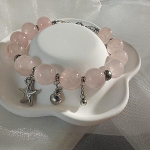 Könnte beinhalten: Ein Rosenquarz-Armband mit Stern-, Muschel- und Perlenanhängern. Das Armband hat große, runde, hellrosa Perlen, die mit silberfarbenen Metallakzenten aufgereiht sind. Das Armband wird auf einer weißen Schale mit gewelltem Rand präsentiert.