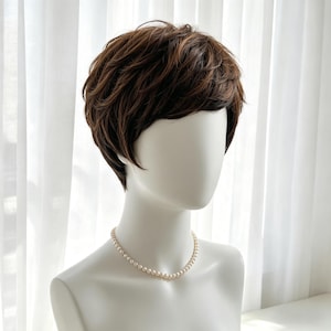 Puede incluir: Una cabeza de maniquí que muestra una peluca corta y castaña con capas. La peluca tiene un aspecto natural con diferentes tonos de marrón. Un delicado collar de perlas adorna el cuello del maniquí, añadiendo un toque de elegancia. El fondo es blanco.