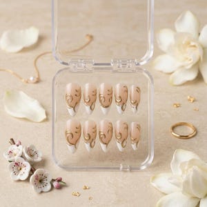 May include: A set of ten almond-shaped press-on nails arranged in a clear plastic storage case. Each nail features a nude base with white tips and intricate gold swirl designs. The nails are displayed in two rows of five within the container.