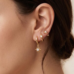 May include: A set of three gold-toned earrings worn in the earlobe and cartilage. The jewelry features a dangling star-shaped charm with a central clear stone, a small hoop with a star accent, and a tiny star-shaped stud, all with a polished finish.