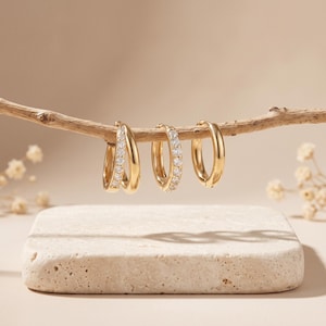 May include: Three gold hoop earrings hanging from a thin wooden branch. The left earring is a double hoop, the middle is a single hoop with embedded clear stones, and the right is a plain gold hoop. All pieces feature a polished metallic finish.