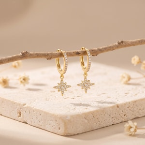 May include: A pair of gold-colored hoop earrings with a starburst charm. The hoops are encrusted with small, clear stones. The starburst charms are also adorned with clear stones. The earrings are displayed on a small branch.