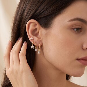 May include: A set of four gold-tone earrings worn in one ear. The top earring is a small leaf-shaped stud with clear stones. Below it are three gold hoop earrings, each featuring a dangling charm: a four-petaled flower, a round stone, and a rectangular stone.