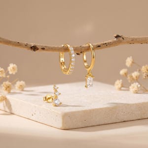 May include: A collection of gold-toned earrings displayed on a neutral background. The earrings include a pair of hoop earrings with clear stones, a pair of hoop earrings with a rectangular pendant, and a stud earring with three clear stones.