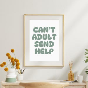 May include: A framed print with the text "CAN'T ADULT SEND HELP" in a light green color. The print is in a light wood frame and is displayed on a desk with a vase of yellow flowers and other decorative items.