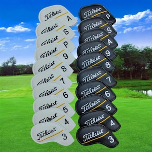 May include: A collection of Titleist golf club head covers. The covers are in two styles: white with yellow accents and black with yellow accents. Each cover is labeled with a club number or letter.