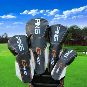 Puede incluir: Un juego de fundas para palos de golf en blanco y negro con la marca "PING" en letras blancas. Las fundas tienen detalles en naranja y el nombre del modelo "G400". El fondo muestra un campo de golf bajo un cielo azul.