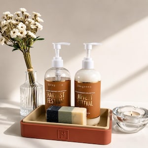 May include: Two clear plastic pump bottles labeled Livegreen Autumn Harvest Collection Harvest Festival, a rectangular bar of soap with three colored segments, and a two-toned terracotta and beige tray, alongside a glass vase with white flowers.
