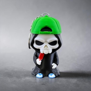 Peut inclure: Un porte-clés représentant une figurine de crâne blanc stylisée portant une casquette de baseball vert vif et une robe à capuche noire. La figurine tient une petite glace rouge à la main et porte des tongs bleu clair, fixée à un anneau.